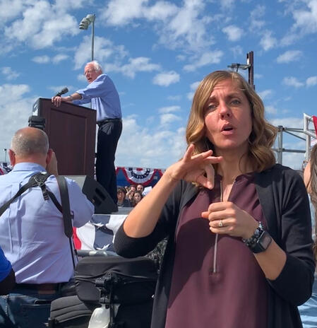 "vote" with Bernie Interpreter outdoors signing vote. Bernie Sanders in the background at a podium.